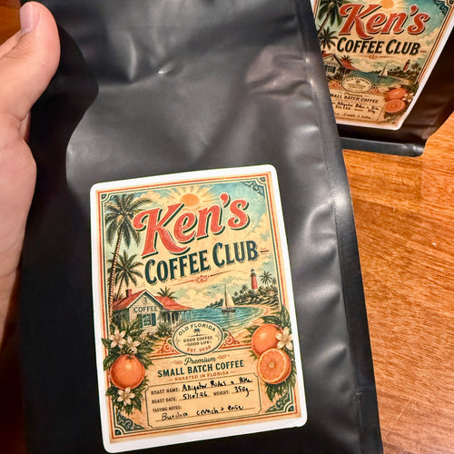 Black coffee bag with 'Ken's Coffee Club' label held by a hand on a wooden surface.